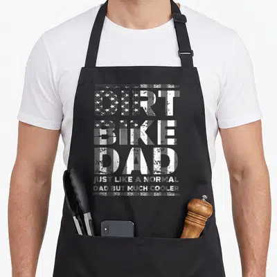 Flag bike apron - Distressed white and grey text reading DIRT BIKE DAD with flag pattern fill on three lines — shown on a black cotton canvas apron against a white background