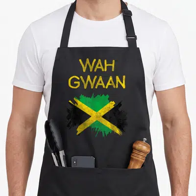 Wah pride apron - Distressed yellow Wah Gwaan text above a green black and yellow paint splatter flag design — shown on a black cotton canvas apron against a white background