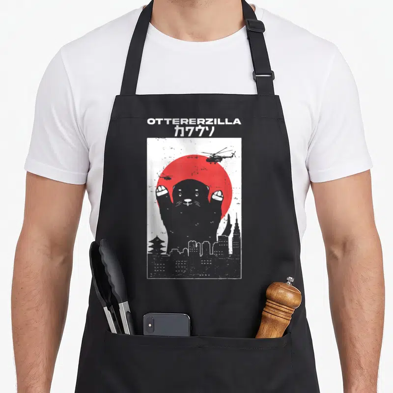Otterzilla otterhound apron - Grey otter silhouette raising paws in front of red sun and city buildings — shown on a black cotton canvas apron against a white background