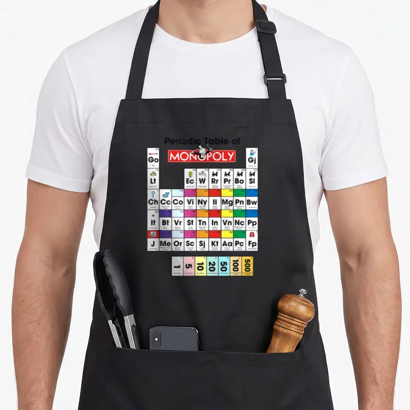 Monopoly table apron - Periodic Table of Monopoly grid with property names, values, and currency bills — shown on a black cotton canvas apron against a white background