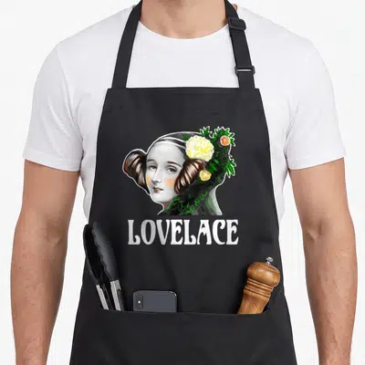 Lovelace ada apron - Realistic woman portrait with green and yellow flowers in hair above white LOVELACE text — shown on a black cotton canvas apron against a white background
