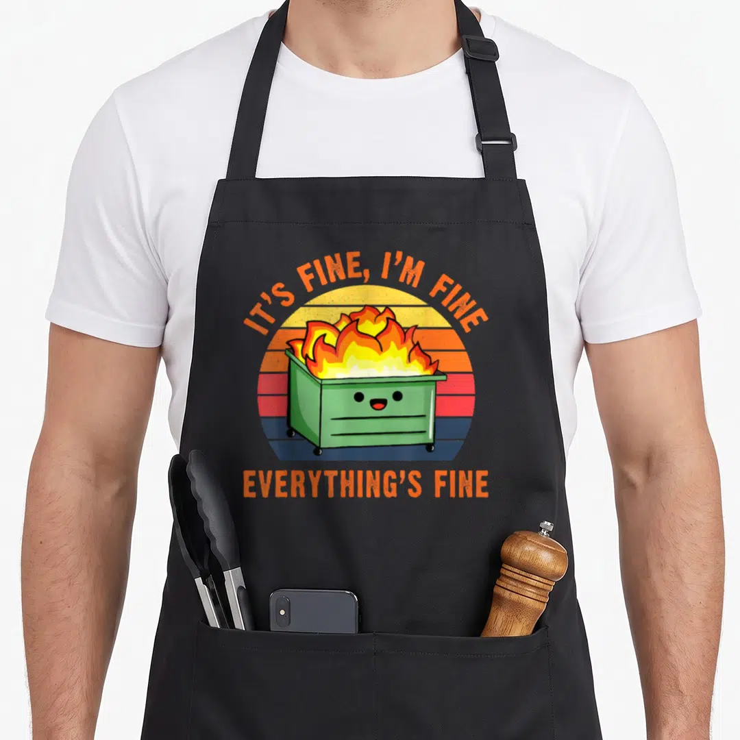 Lil its fine apron - Cartoon green dumpster on fire with smiling face and orange text over retro striped sun — shown on a black cotton canvas apron against a white background
