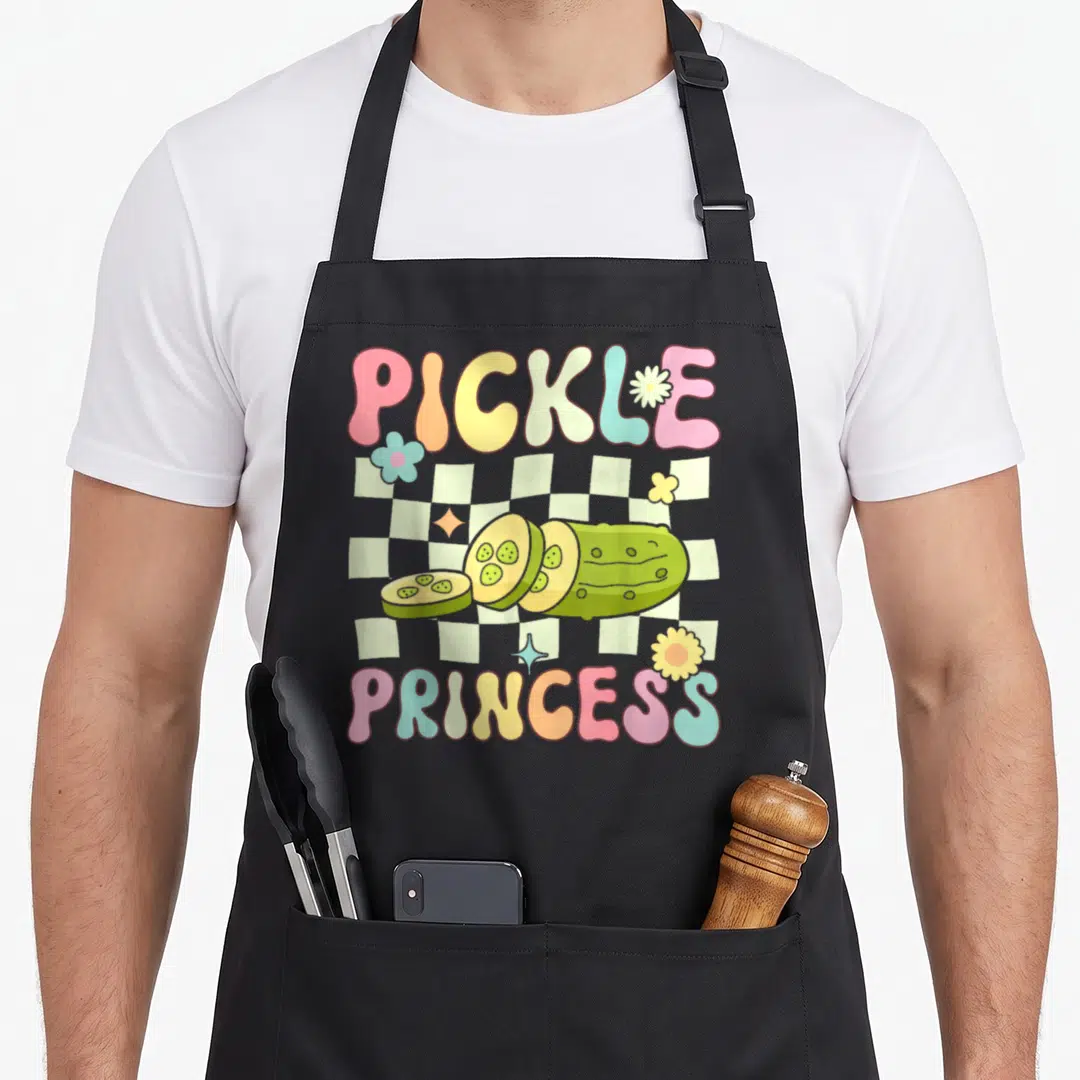Pickle princess apron - Cartoon sliced pickle on checkerboard with PICKLE PRINCESS text and pastel flowers — shown on a black cotton canvas apron against a white background