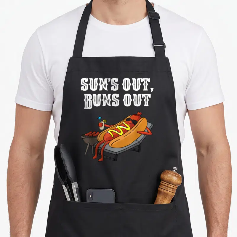 Sun sausage apron - Cartoon hot dog on lounge chair with sunglasses and text SUN'S OUT BUNS OUT — shown on a black cotton canvas apron against a white background