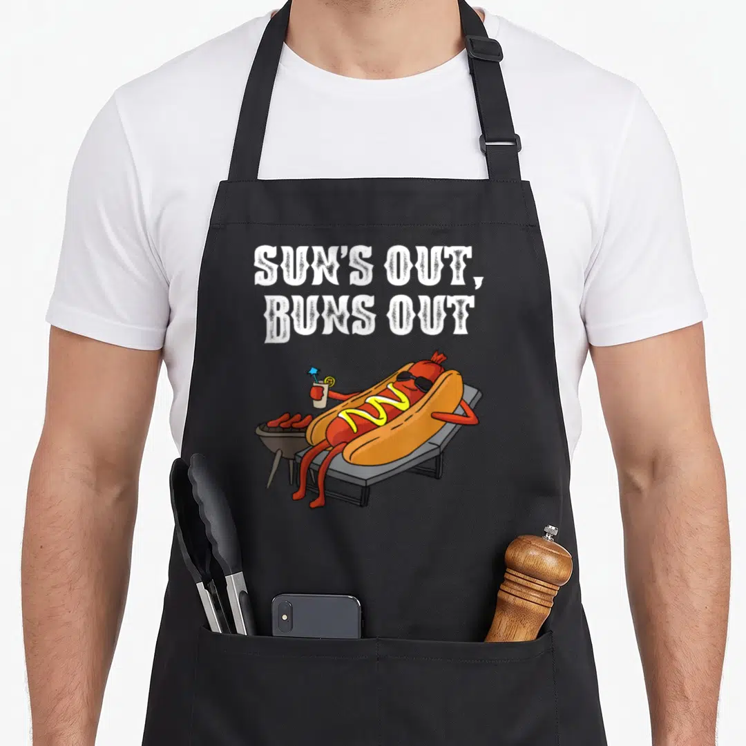 Sun sausage apron - Cartoon hot dog on lounge chair with sunglasses and text SUN'S OUT BUNS OUT — shown on a black cotton canvas apron against a white background