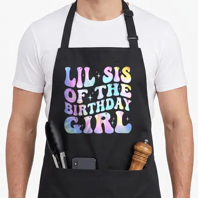 Sister white lily apron - Tie-dye typography reading LIL SIS OF THE BIRTHDAY GIRL with sparkles — shown on a black cotton canvas apron against a white background
