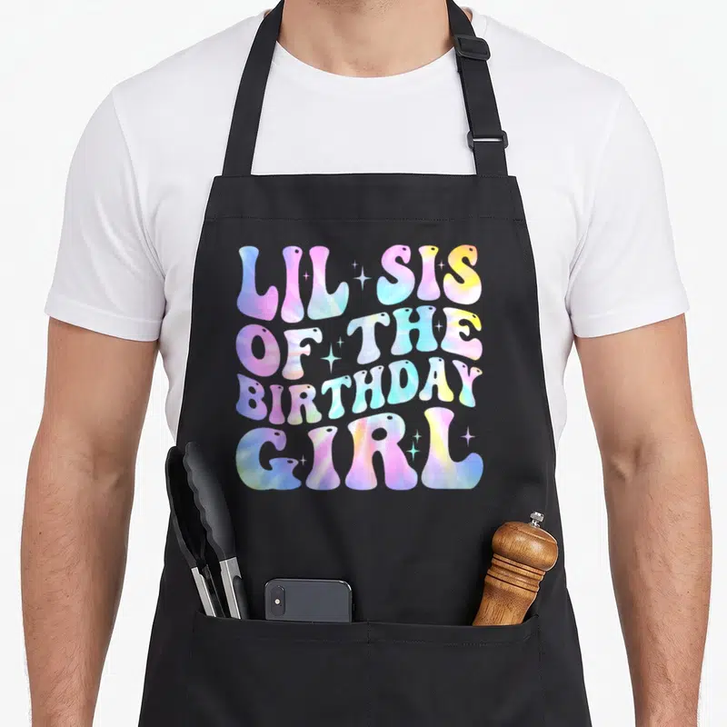 Sister white lily apron - Tie-dye typography reading LIL SIS OF THE BIRTHDAY GIRL with sparkles — shown on a black cotton canvas apron against a white background