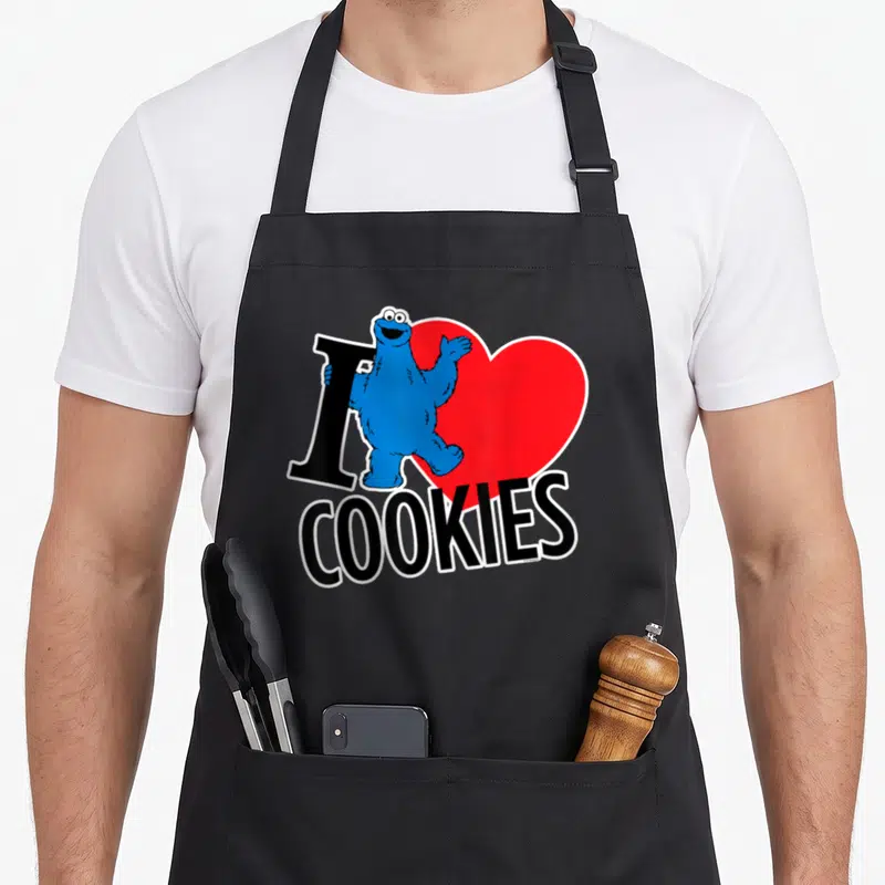 Funny cookie cookies apron - Blue character standing over red heart next to black I and COOKIES text — shown on a black cotton canvas apron against a white background