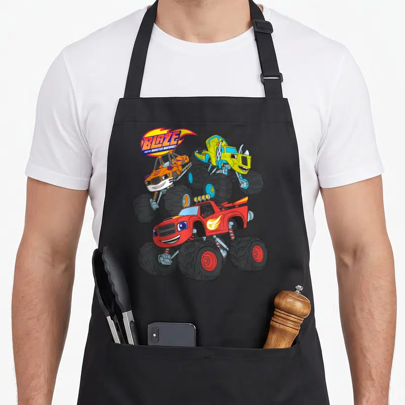 Stripe machine apron - Blaze logo and three monster trucks in red, yellow, and orange cartoon style — shown on a black cotton canvas apron against a white background