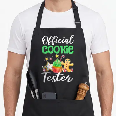 Cute holiday baking apron - Official COOKIE Tester text surrounding a cupcake, gingerbread man, star cookie, and candy — shown on a black cotton canvas apron against a white background