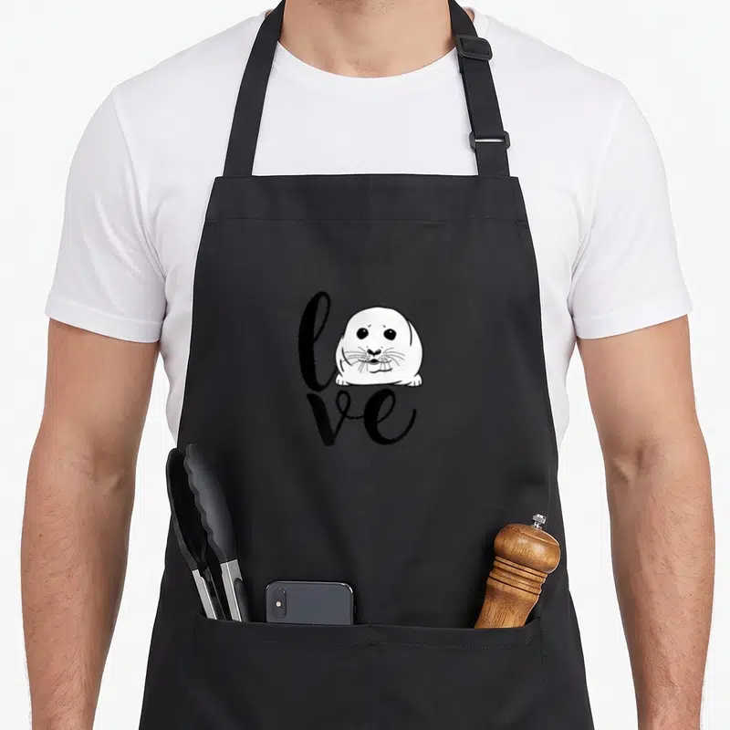 Baby harp apron - Cartoon seal head replacing the letter O in the word love printed in black script — shown on a black cotton canvas apron against a white background