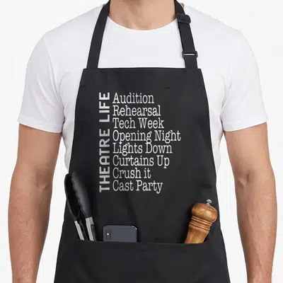 Broadway actor theatre apron - Vertical THEATRE LIFE text beside a list of eight production phrases in white font — shown on a black cotton canvas apron against a white background
