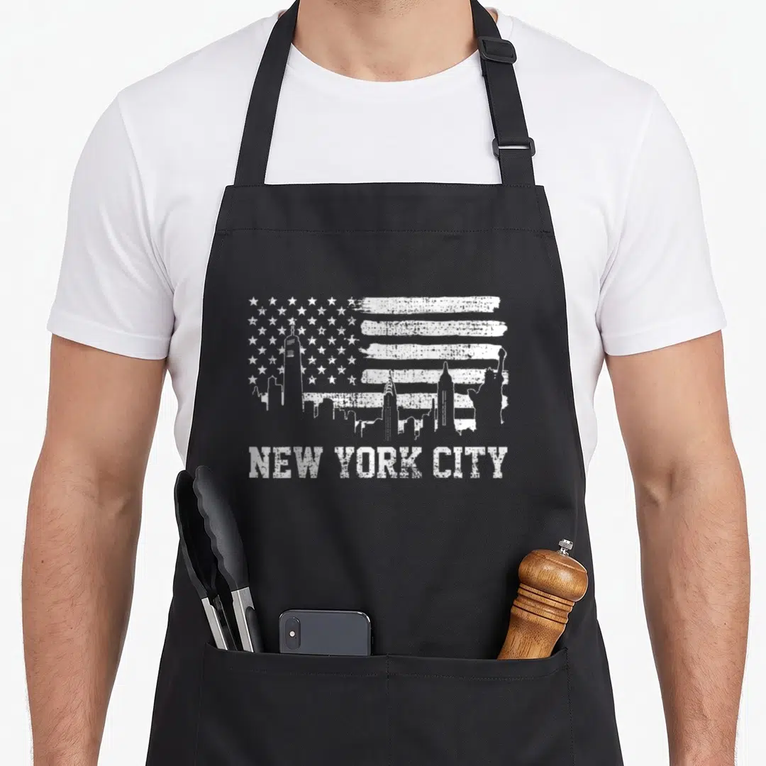 York apple apron - White distressed New York City skyline inside a US flag pattern with text below — shown on a black cotton canvas apron against a white background