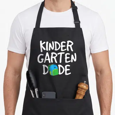 Kinder dude apron - Stacked white text KINDER GARTEN DADE with blue and green backpack replacing the letter — shown on a black cotton canvas apron against a white background
