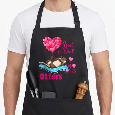 Adorable balloon apron - Cartoon otter floating in blue water holding pink heart balloons next to pink text — shown on a black cotton canvas apron against a white background