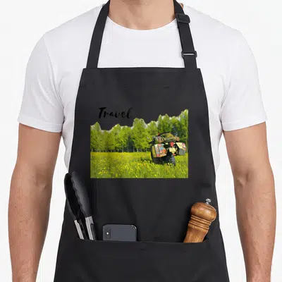 Adventure moto bike apron - Photo of loaded motorcycle in yellow flowers with green trees and Travel text — shown on a black cotton canvas apron against a white background
