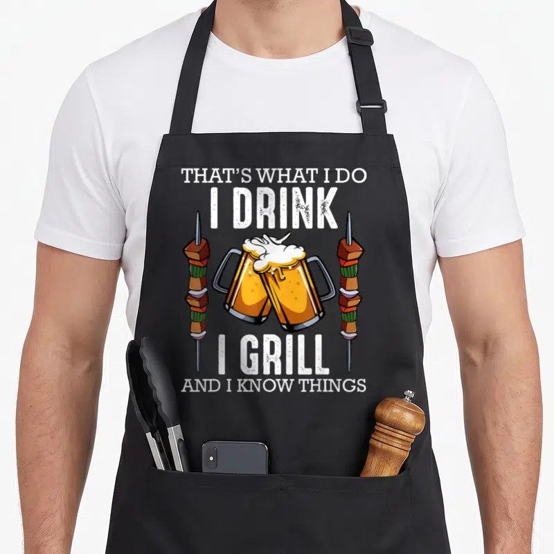 Drink speck apron - Two beer mugs toasting between vertical shish kebabs with stacked white text above and below — shown on a black cotton canvas apron against a white background