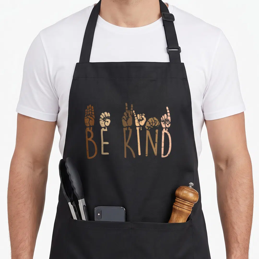 Hand asl sign for apron - Six hands in brown beige tan and peach rising from the words BE KIND — shown on a black cotton canvas apron against a white background