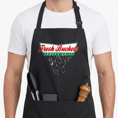 Basketball bucket apron - Fresh Buckets Served Daily banner over basketball net in red green and black — shown on a black cotton canvas apron against a white background