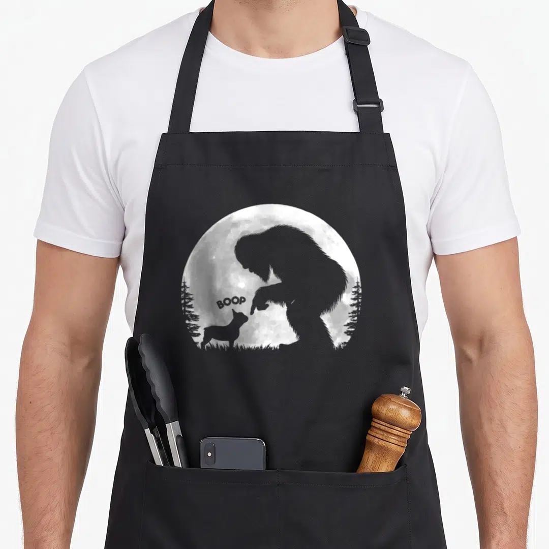 Hand french bulldog apron - Bigfoot silhouette booping a dog nose with BOOP text inside a moon circle — shown on a black cotton canvas apron against a white background