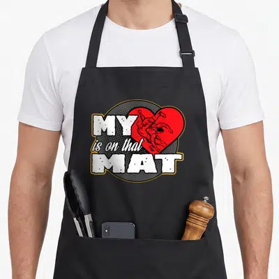 Mat mama apron - MY is on that MAT text beside red heart with two cartoon wrestlers on grey halftone circle — shown on a black cotton canvas apron against a white background