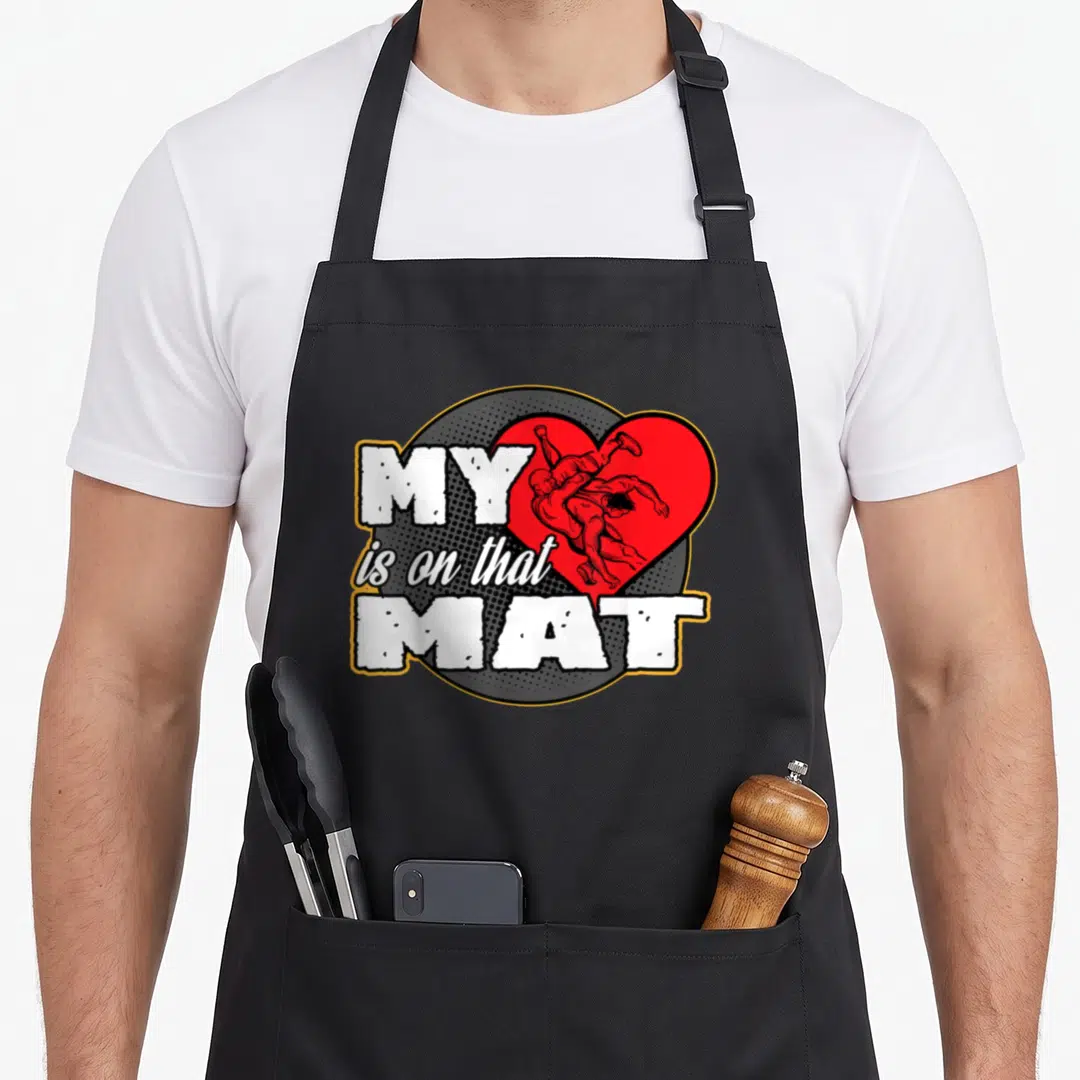 Mat mama apron - MY is on that MAT text beside red heart with two cartoon wrestlers on grey halftone circle — shown on a black cotton canvas apron against a white background