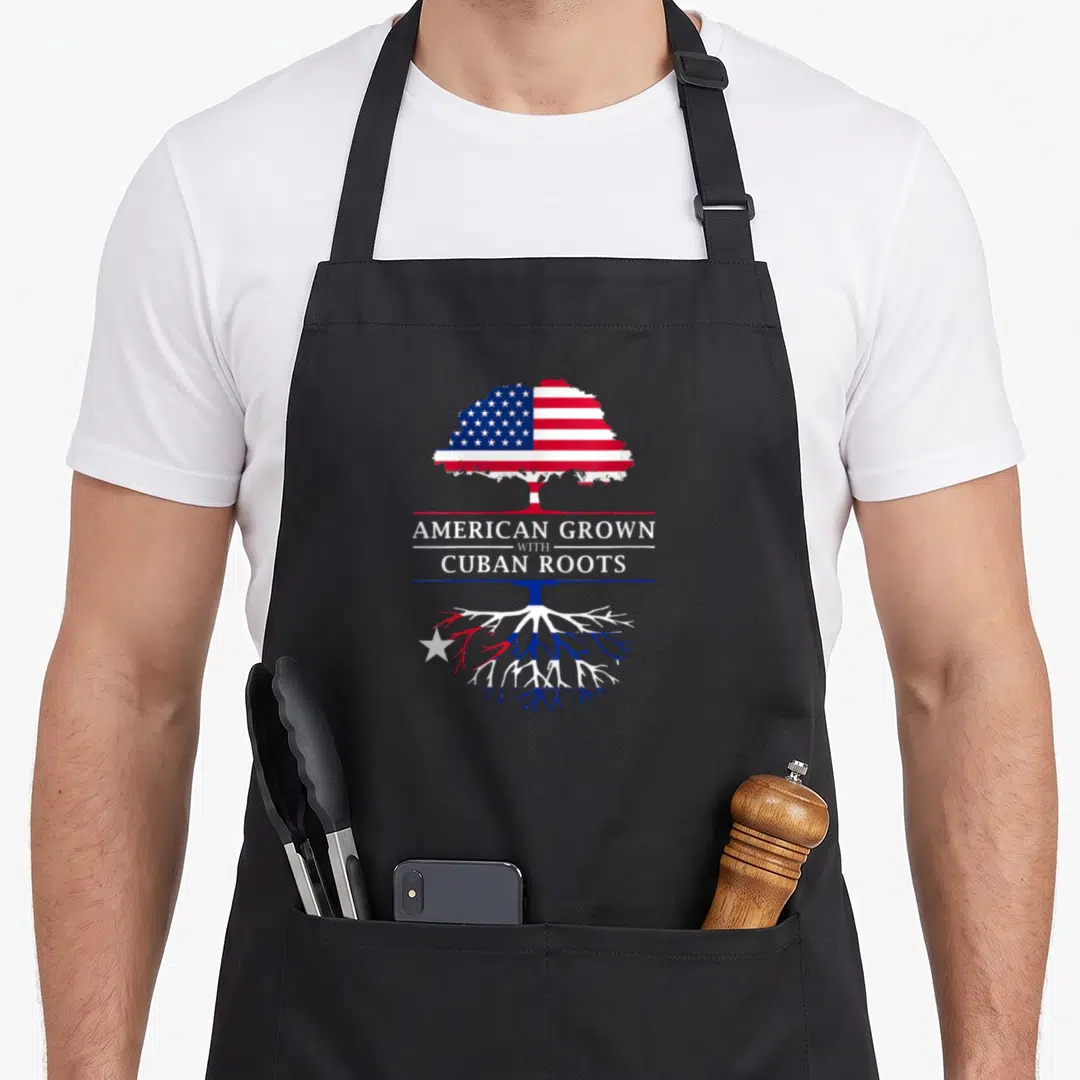 Root cuban apron - Tree graphic with US flag leaves and Cuban flag roots separating stacked text — shown on a black cotton canvas apron against a white background