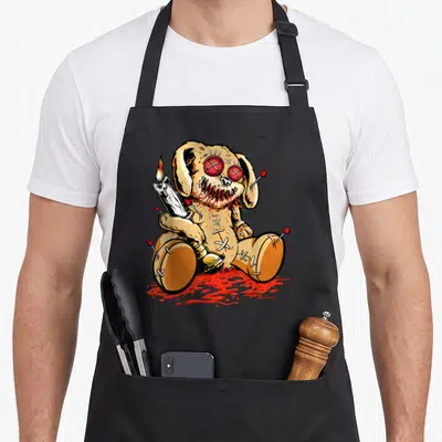 Voodoo teddy bear apron - Tan cartoon voodoo doll with red pins holding white candle in red pool — shown on a black cotton canvas apron against a white background