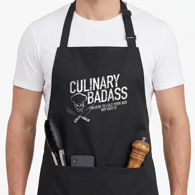 Feed culinary badass apron - White distressed skull in chef hat with crossed knife and fork and bold text — shown on a black cotton canvas apron against a white background