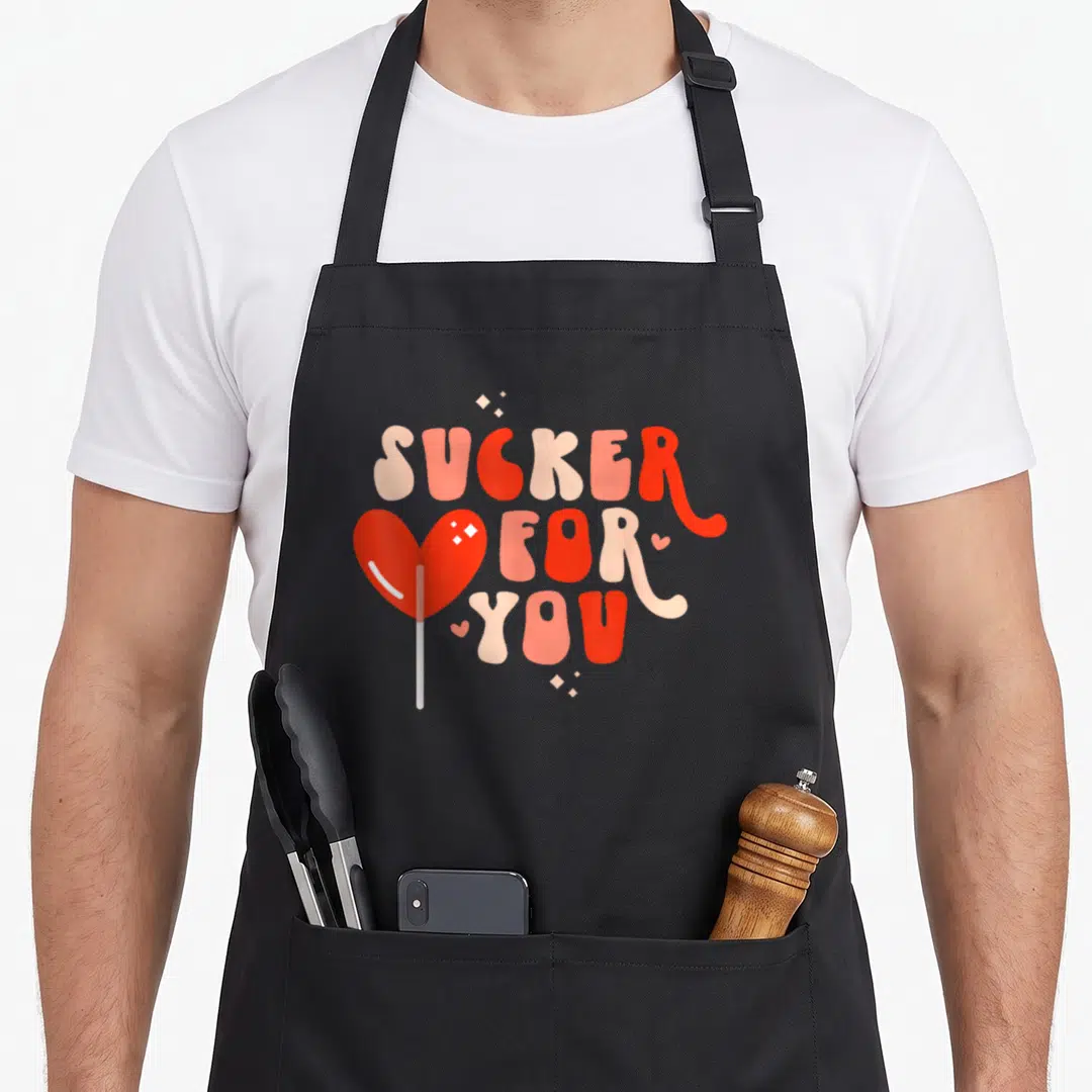 Sucker cotton candy apron - Heart lollipop with SUCKER FOR YOU text in red pink white — shown on a black cotton canvas apron against a white background