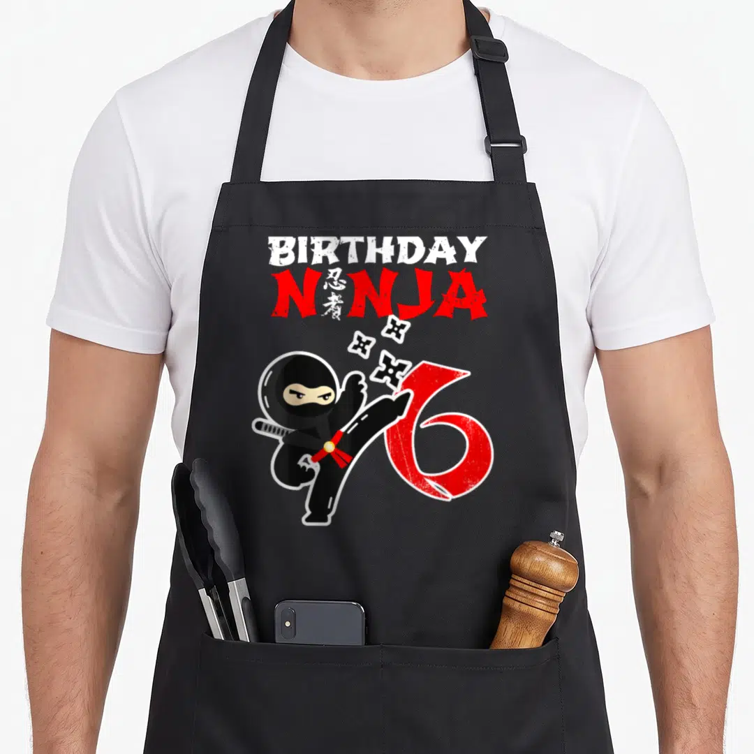 Favour birthday apron - Cartoon ninja kicking red number 6 with BIRTHDAY NINJA text and flying shuriken — shown on a black cotton canvas apron against a white background