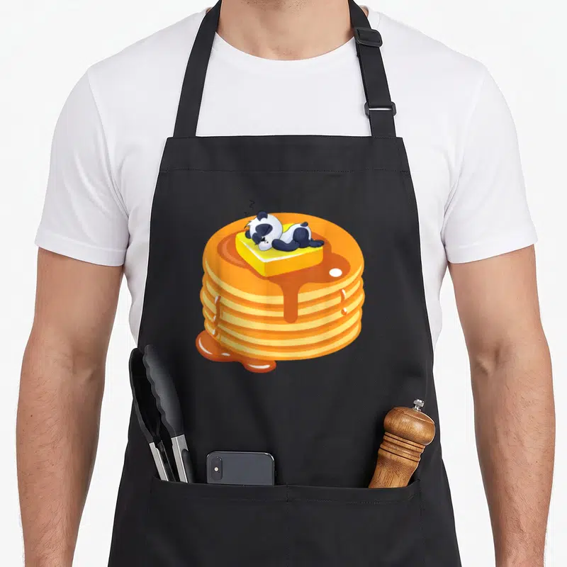 Baby pancake apron - Cartoon panda sleeping on butter atop five pancakes with dripping orange syrup — shown on a black cotton canvas apron against a white background