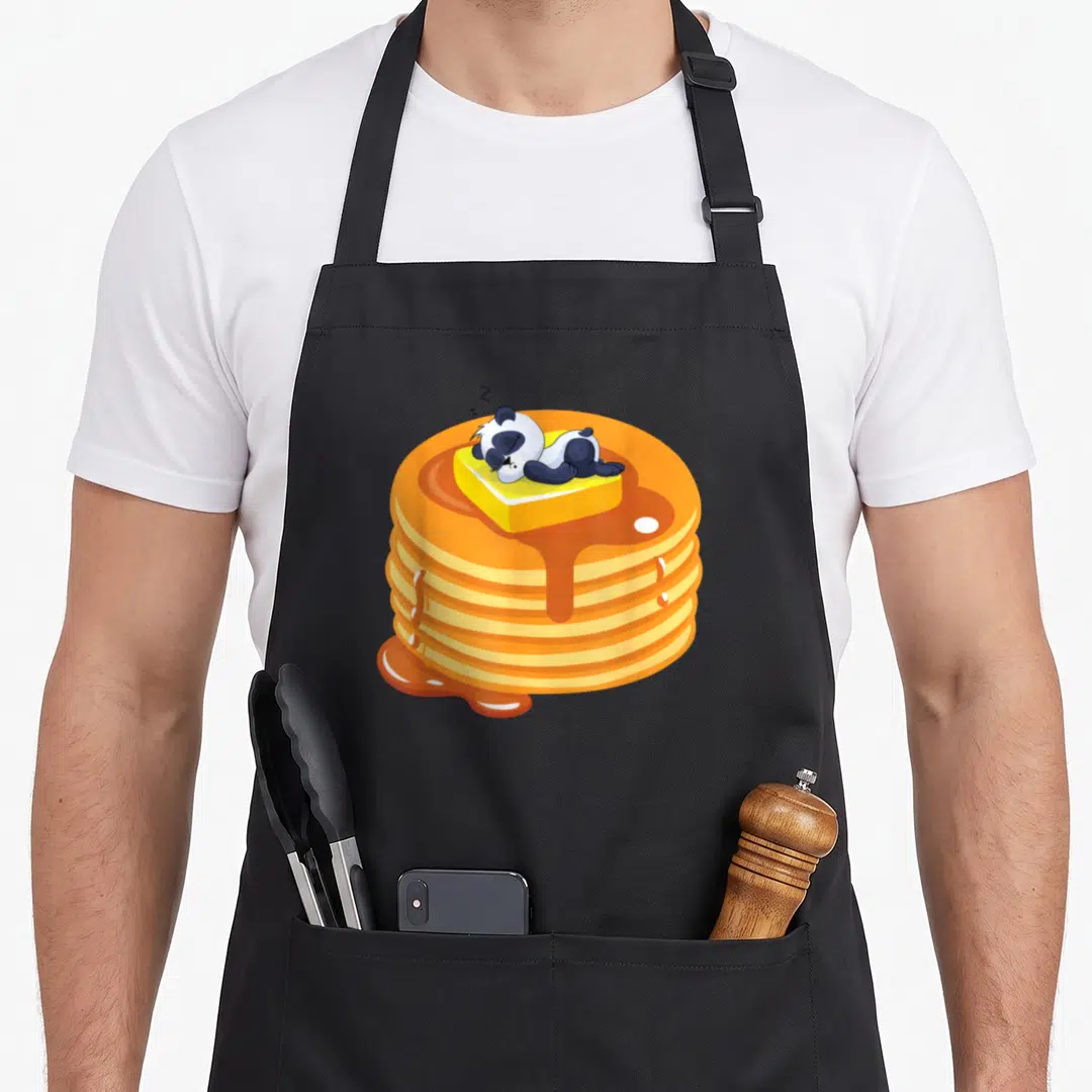 Baby pancake apron - Cartoon panda sleeping on butter atop five pancakes with dripping orange syrup — shown on a black cotton canvas apron against a white background