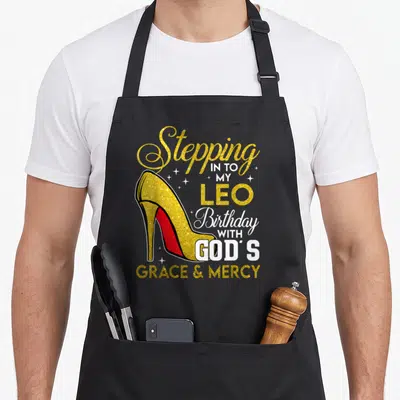 Stepping leo birthday apron - Gold glitter high heel shoe with red sole next to stacked birthday text and small stars — shown on a black cotton canvas apron against a white background