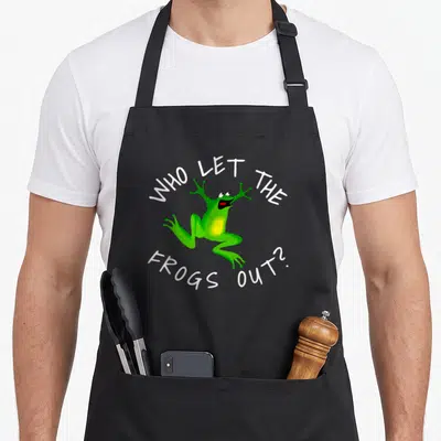 Frog passover apron - Green cartoon frog with arched white text WHO LET THE FROGS OUT? — shown on a black cotton canvas apron against a white background