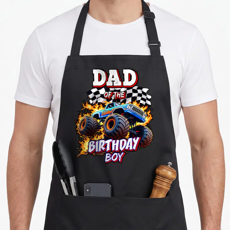 Retro birthday truck apron - DAD OF THE BIRTHDAY BOY text over blue monster truck with flames and checkered flag — shown on a black cotton canvas apron against a white background