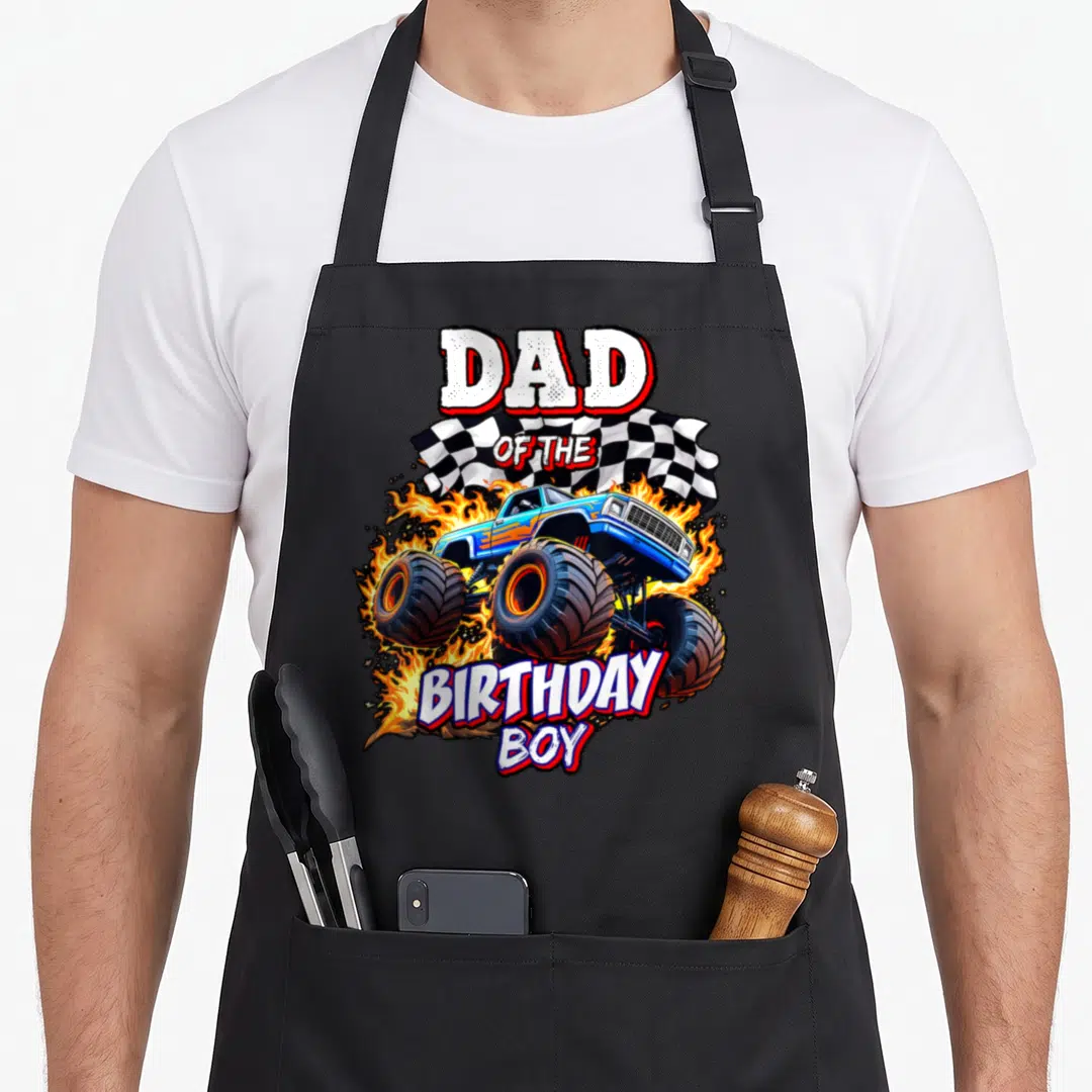 Retro birthday truck apron - DAD OF THE BIRTHDAY BOY text over blue monster truck with flames and checkered flag — shown on a black cotton canvas apron against a white background