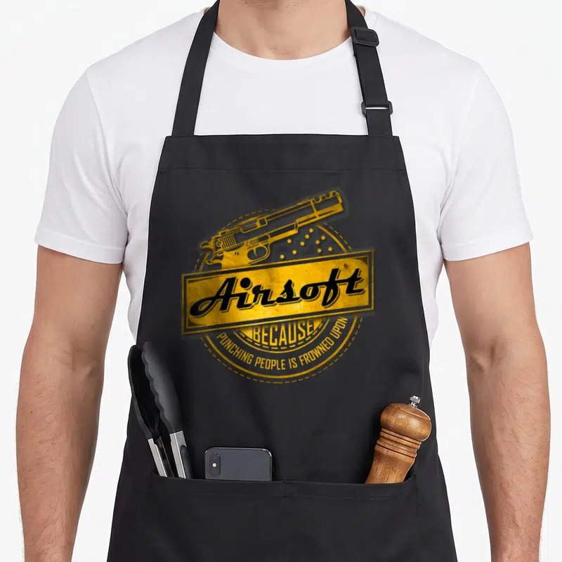 Punching airsoft apron - Yellow retro pistol graphic with Airsoft text and curved slogan on circular background — shown on a black cotton canvas apron against a white background