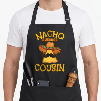 Cousin mexican apron - Cartoon nacho chip in sombrero with mustache and stacked yellow text NACHO AVERAGE COUSIN — shown on a black cotton canvas apron against a white background