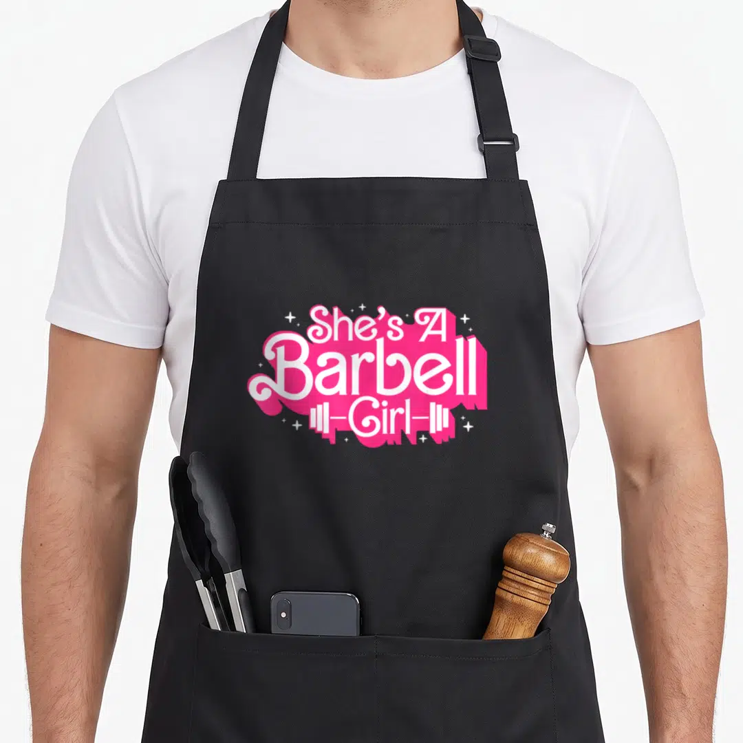 Barbell weightlifter apron - She's A Barbell -Girl- text in white over pink splatter with stars and barbell icon — shown on a black cotton canvas apron against a white background