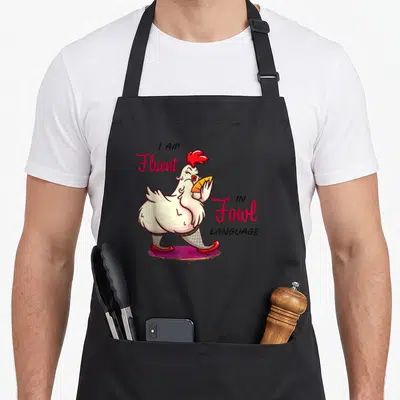 Chicken fowl language apron - Cartoon white chicken holding fan with text I AM Fluent IN Fowl LANGUAGE — shown on a black cotton canvas apron against a white background
