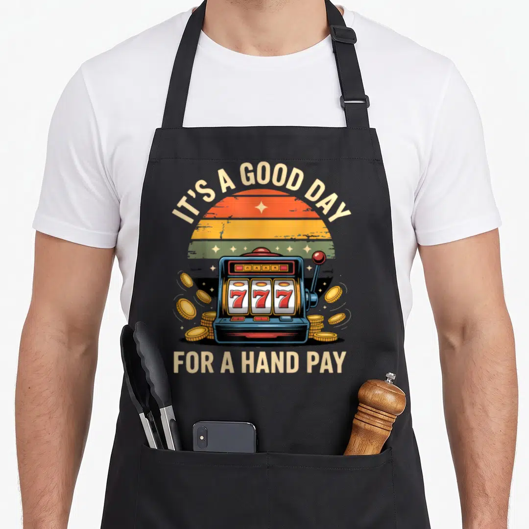 Hand casino aprons - Retro slot machine with 777 reels, gold coins, and striped sun behind arched text — shown on a black cotton canvas apron against a white background