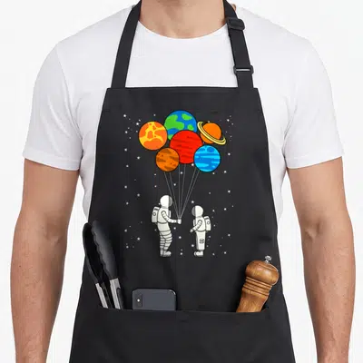 Galaxy balloon apron - Two astronauts holding strings attached to a cluster of colorful planet balloons among white — shown on a black cotton canvas apron against a white background