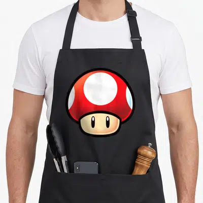 Face mushroom apron - Cartoon red mushroom with white spots and black outline centered on grey background — shown on a black cotton canvas apron against a white background