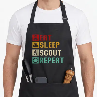 Eat unique apron - Four colored squares with icons next to Eat Sleep Scout Repeat text — shown on a black cotton canvas apron against a white background