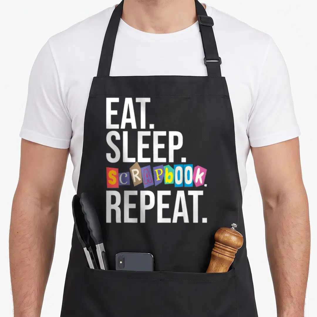 Eat scrapbooking apron - EAT SLEEP SCRAPBOOK REPEAT text with colorful paper scraps stacked vertically — shown on a black cotton canvas apron against a white background