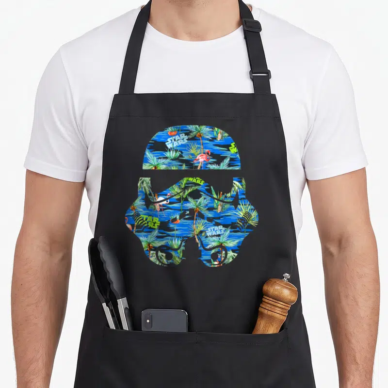 Helmet hawaiian apron - Stormtrooper helmet silhouette filled with palm trees, flamingos, and scattered STAR WARS text — shown on a black cotton canvas apron against a white background