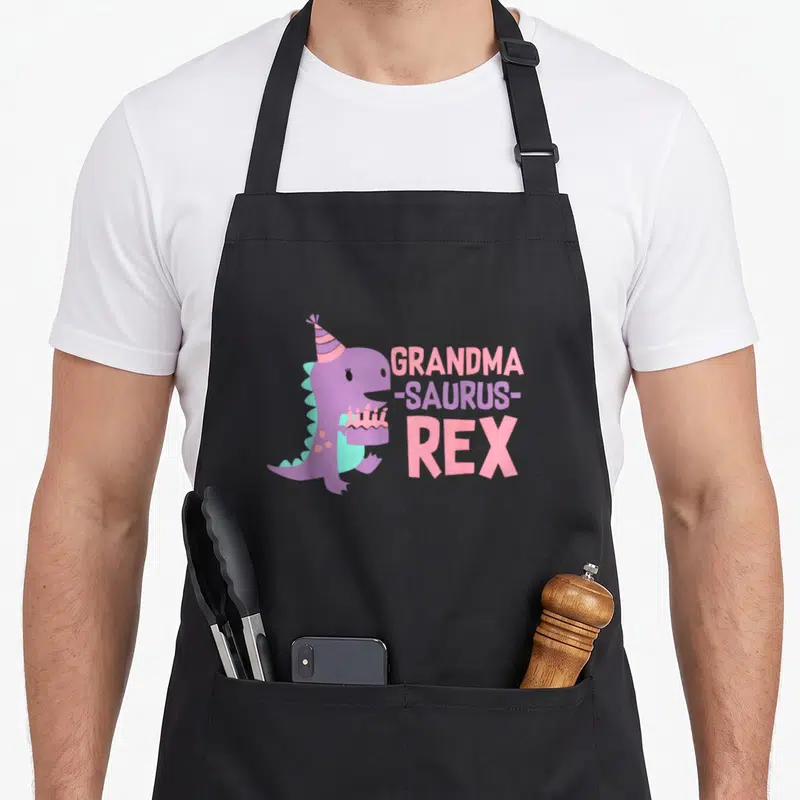 Dino grandma in apron - Purple cartoon dinosaur with teal spikes holding a pink cake next to stacked text — shown on a black cotton canvas apron against a white background