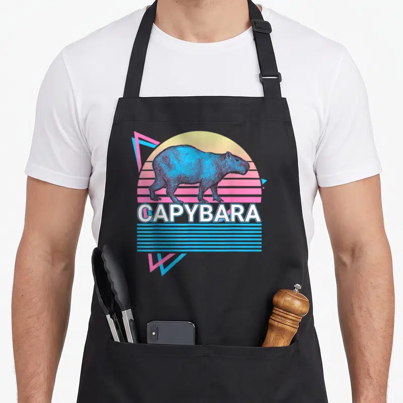 Funny capybara apron - Blue capybara silhouette on yellow pink gradient sun with geometric triangles and CAPYBARA text — shown on a black cotton canvas apron against a white background