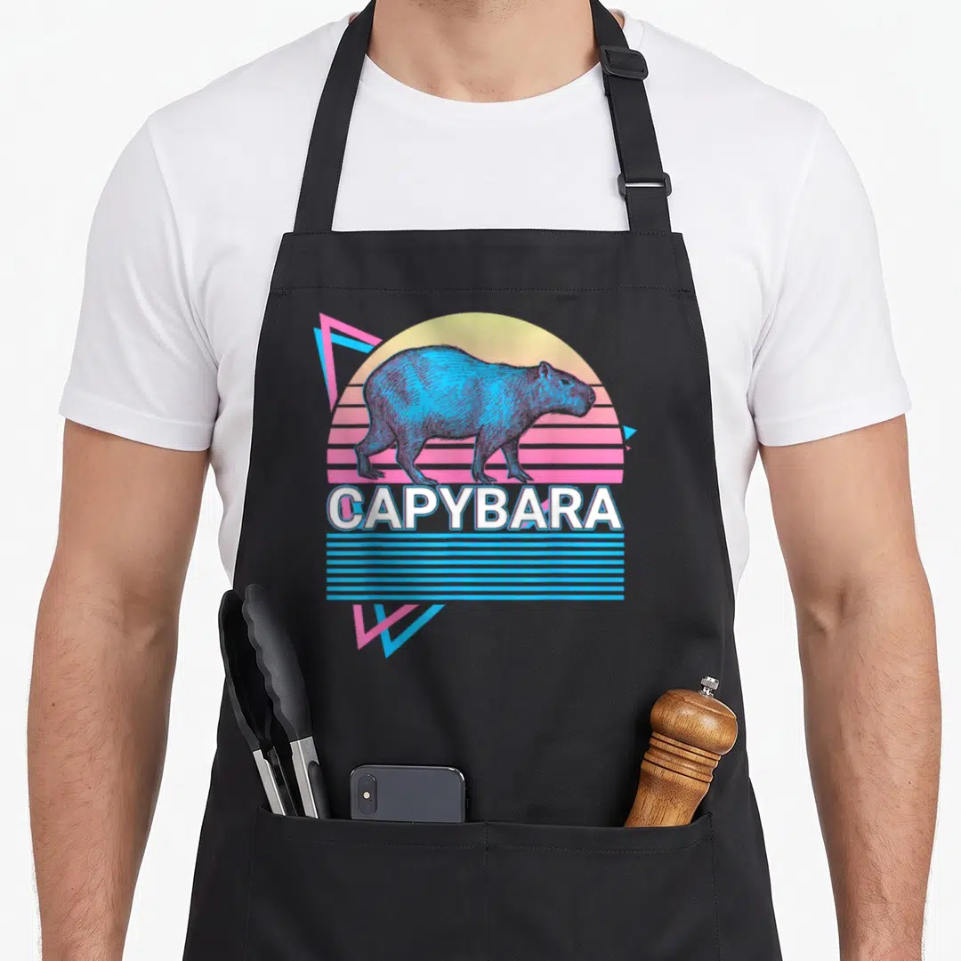 Funny capybara apron - Blue capybara silhouette on yellow pink gradient sun with geometric triangles and CAPYBARA text — shown on a black cotton canvas apron against a white background
